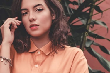 Person with curled shoulder-length brown hair wearing a collared orange top has their right arm bent and slightly raised near right cheek to show a bracelet on their wrist