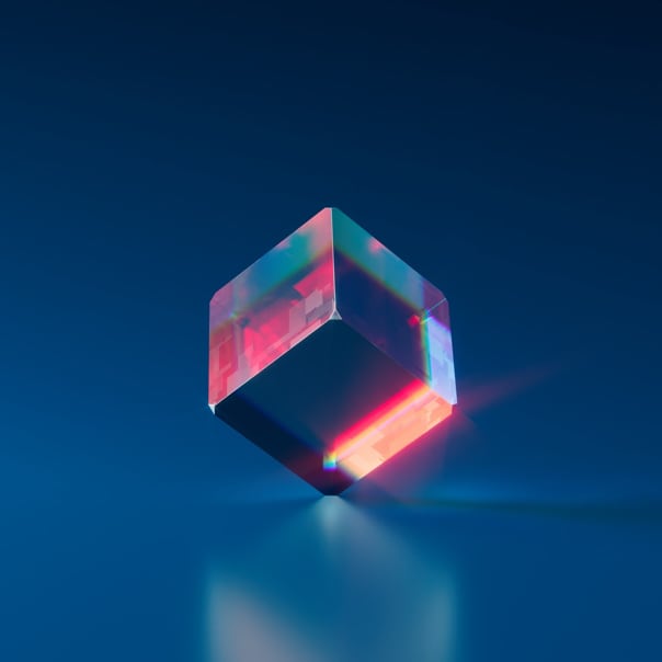 A glass cube against a blue backdrop, balanced on one tip angled to show the bottom side and its faces partially reflecting light in reddish hues